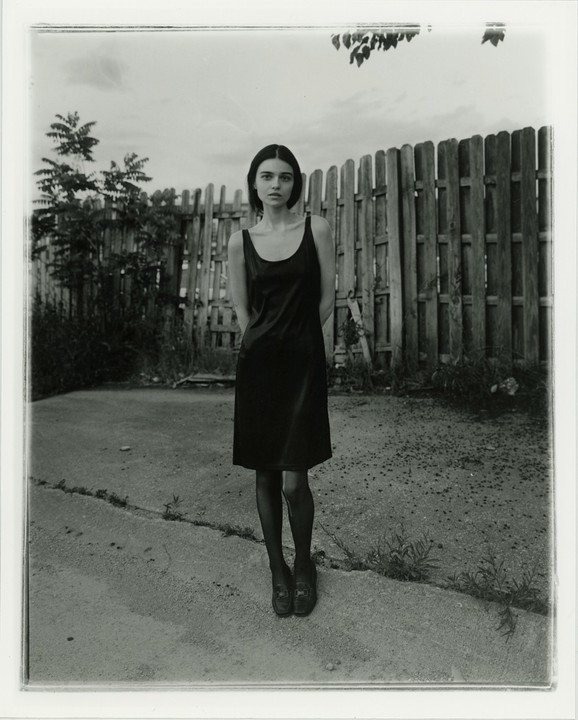 A woman in a dark dress stands in front of a wooden fence with trees and a cloudy sky in the background.