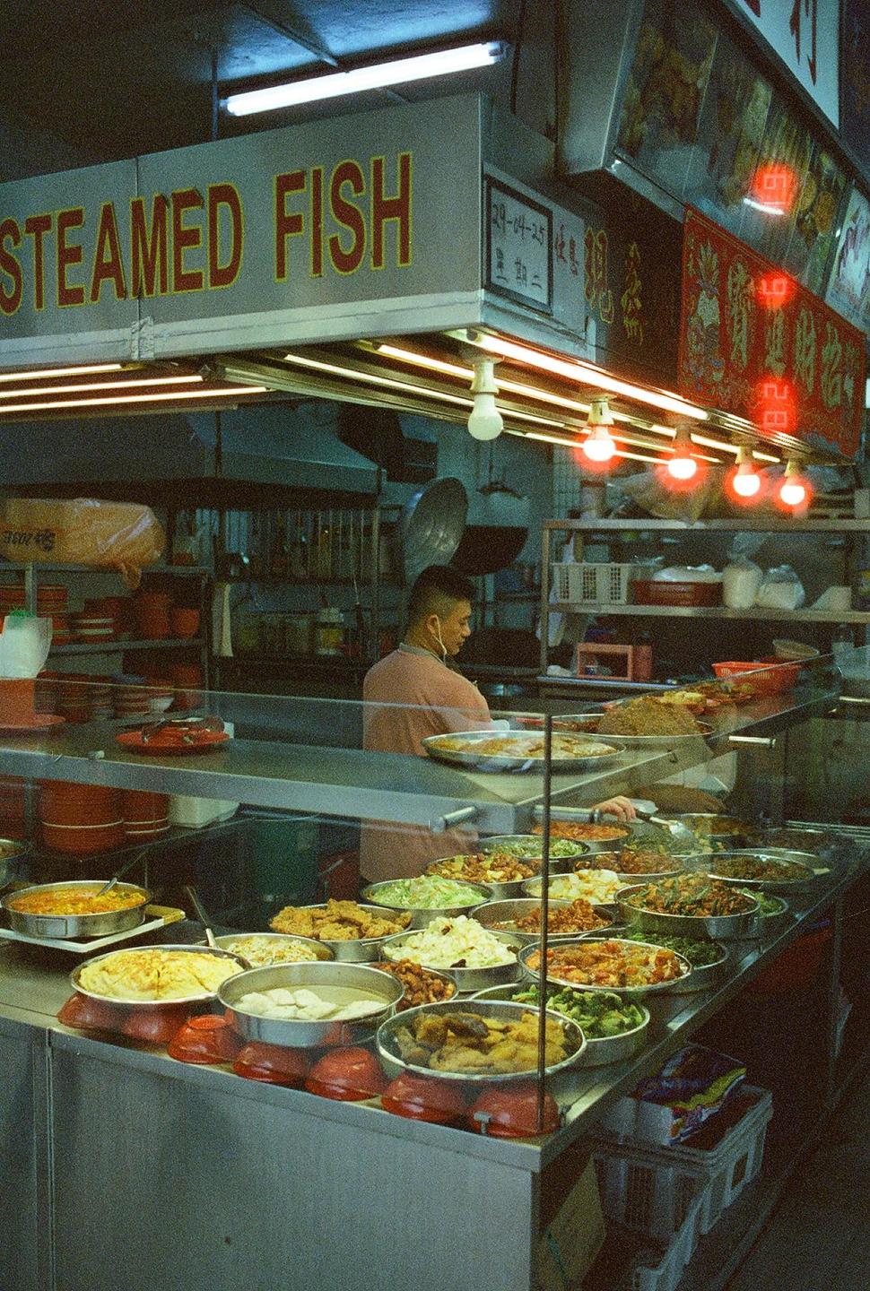 A man stands behind a counter filled with various dishes in a food stall.