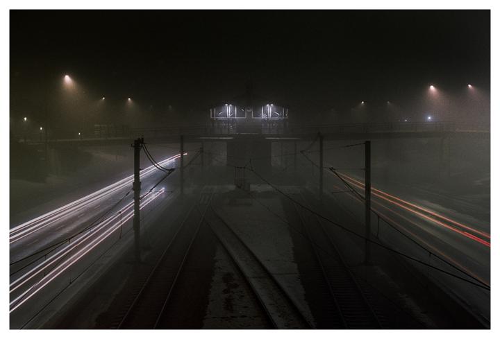 Light trails from vehicles and a train station are visible through the fog at night.