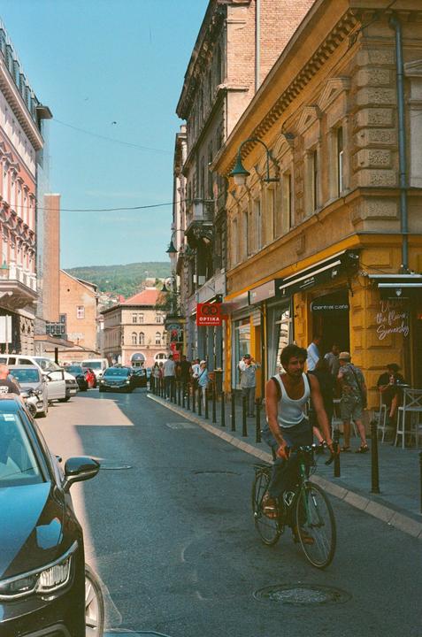 A man rides a bicycle down a street lined with buildings and parked cars, with people walking on the sidewalk.