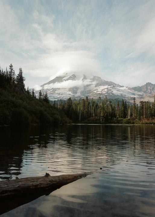 A snow-capped mountain with clouds around its peak rises above a forest and a lake with a log in the foreground.