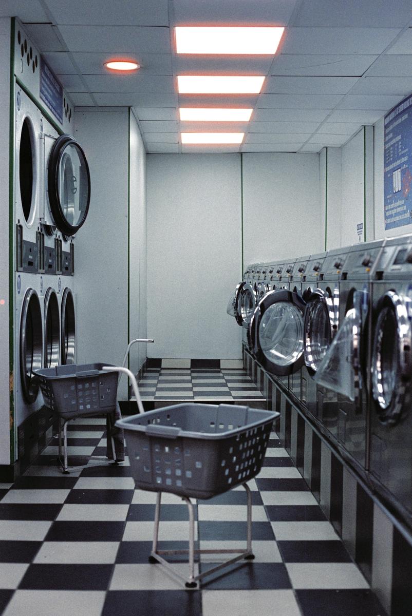 A long, narrow laundromat with a checkered floor is lined with washing machines and laundry baskets.