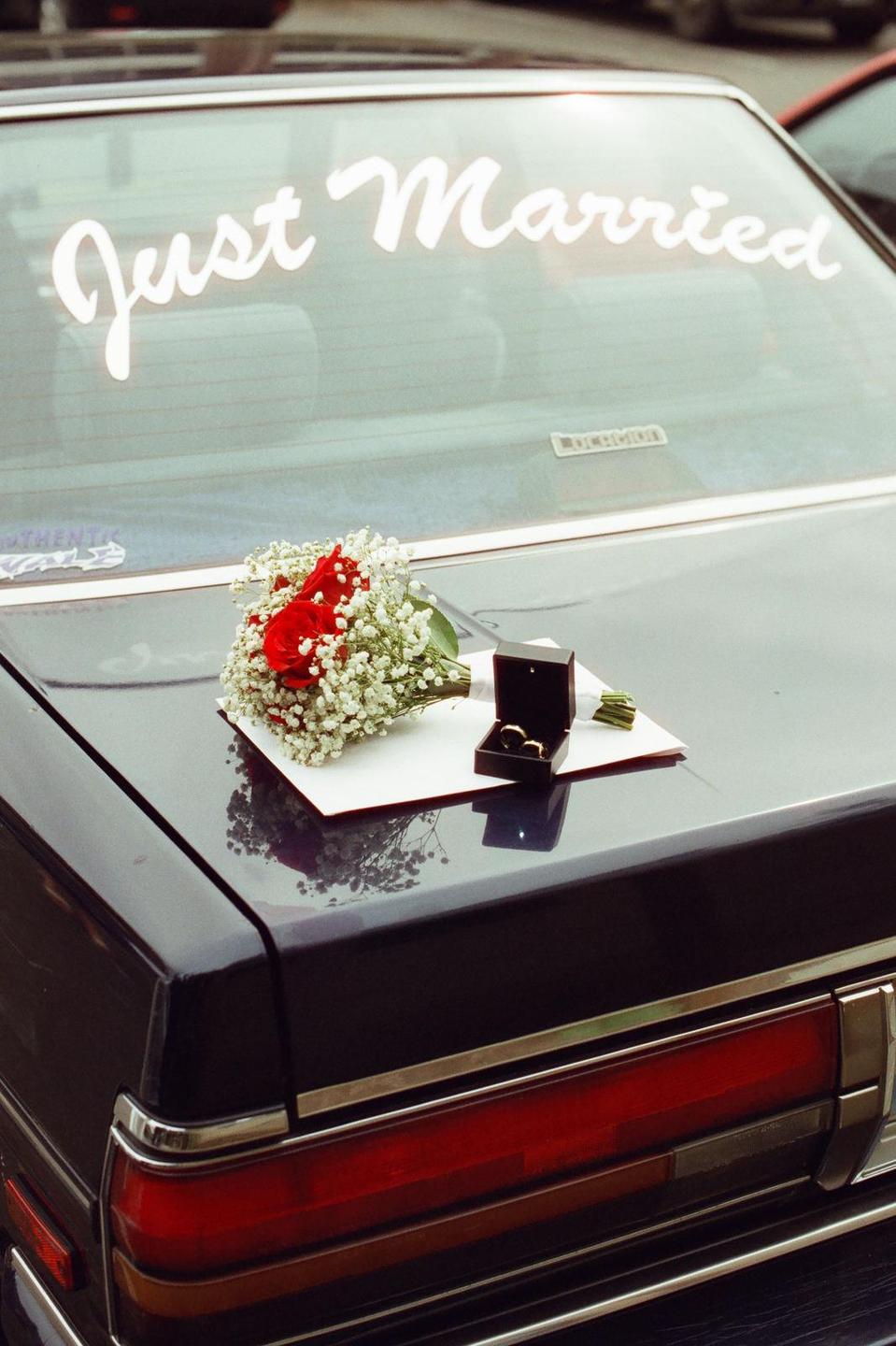 A bouquet of red roses and baby's breath, a ring box with two rings, and a white card sit on the trunk of a car with a "Just Married" sign on the back window.