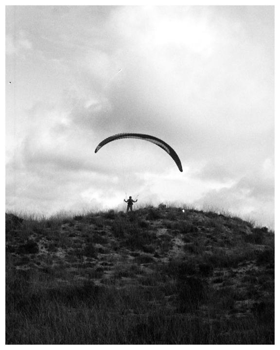 A person stands on a grassy hill with a paraglider above them under a cloudy sky.