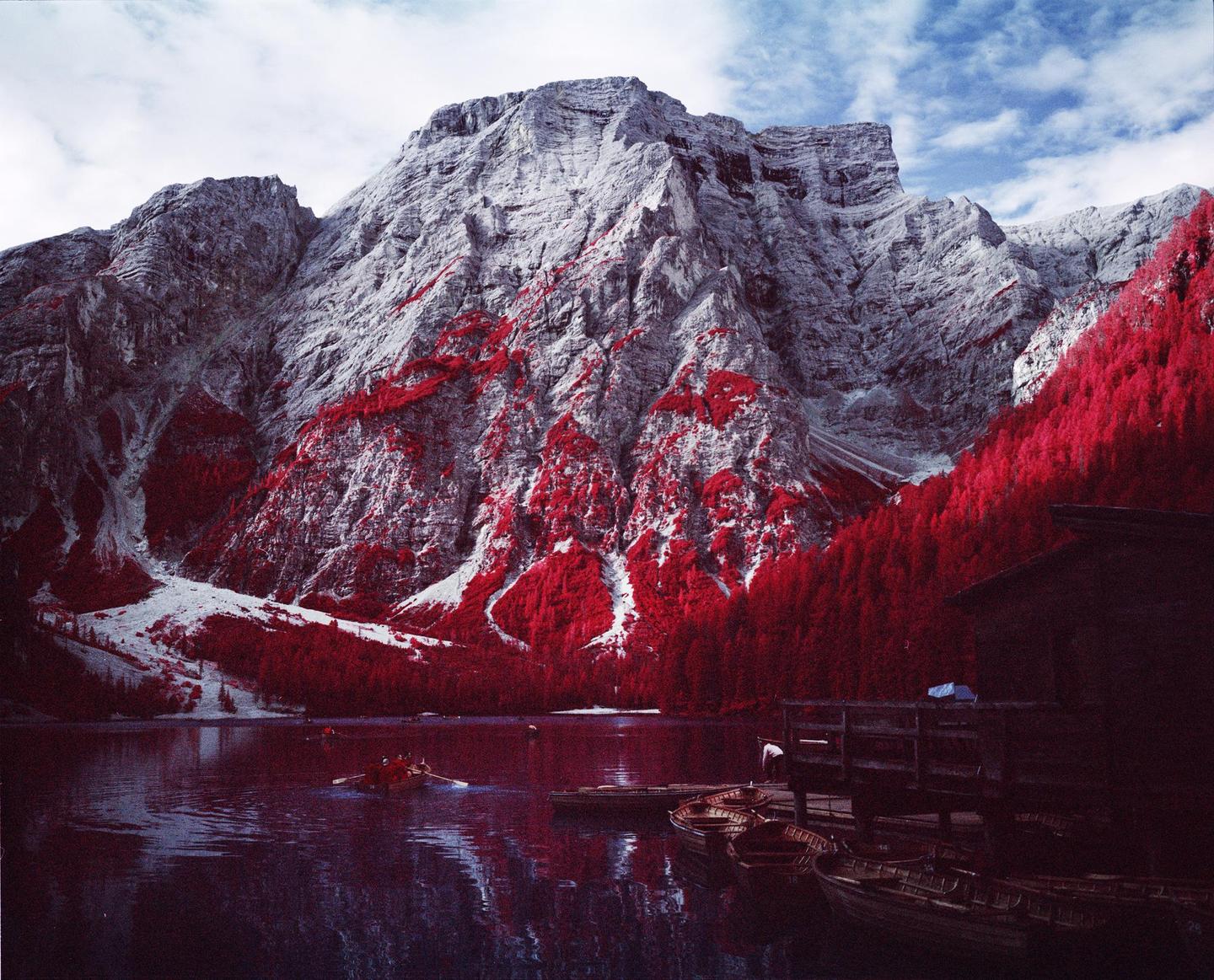 A lake with boats and a boathouse is surrounded by mountains and red trees under a cloudy sky.