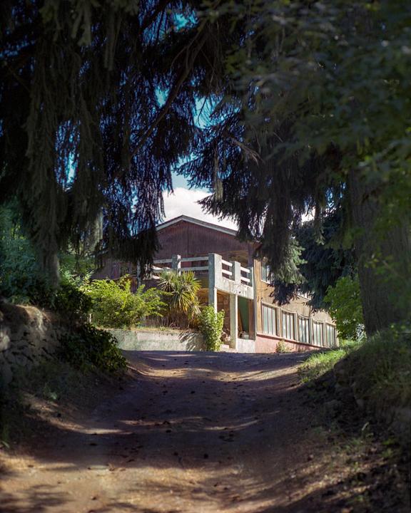 A dirt road leads to a house with a balcony, framed by dark trees.