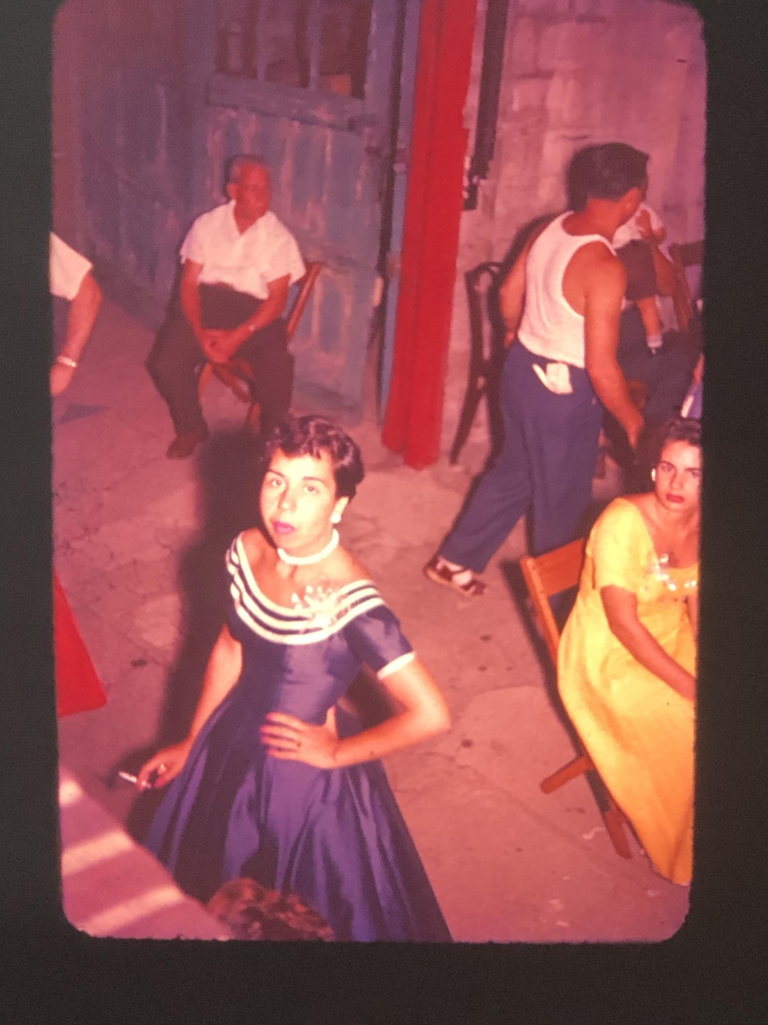 A woman in a blue dress holds a cigarette while standing among other people in an outdoor setting.