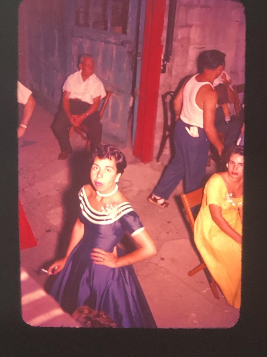 A woman in a blue dress holds a cigarette while standing among other people in an outdoor setting.