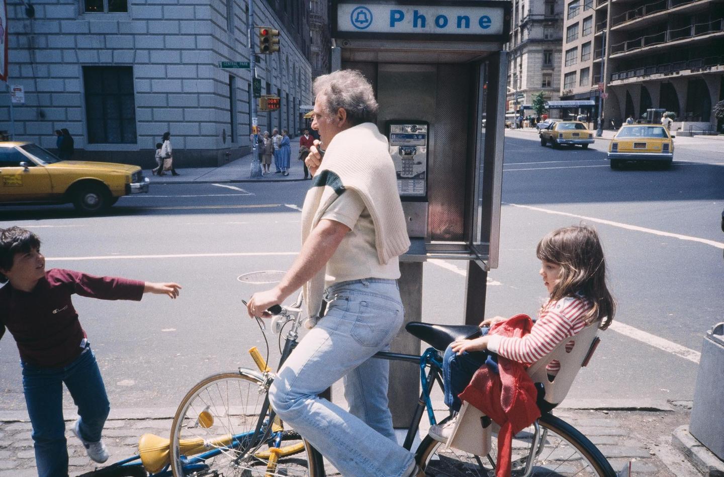A man on a bicycle with two children stands next to a payphone booth on a city street with yellow taxis.