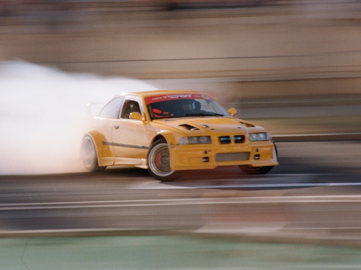 A yellow race car drifts on a track, creating a large cloud of smoke from its tires.
