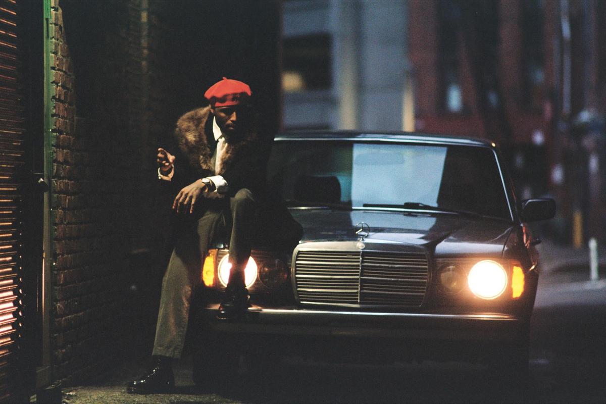 A man in a red beret and fur-collared coat sits on the hood of a car with its headlights on at night.