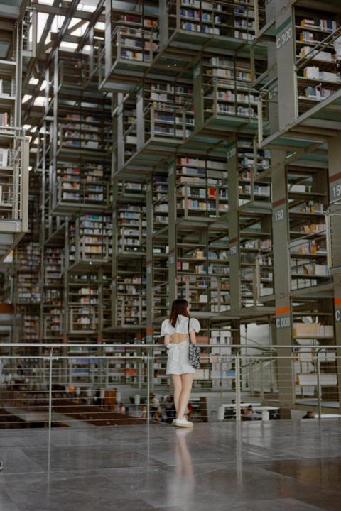 A woman stands on a balcony overlooking a vast library with towering bookshelves.