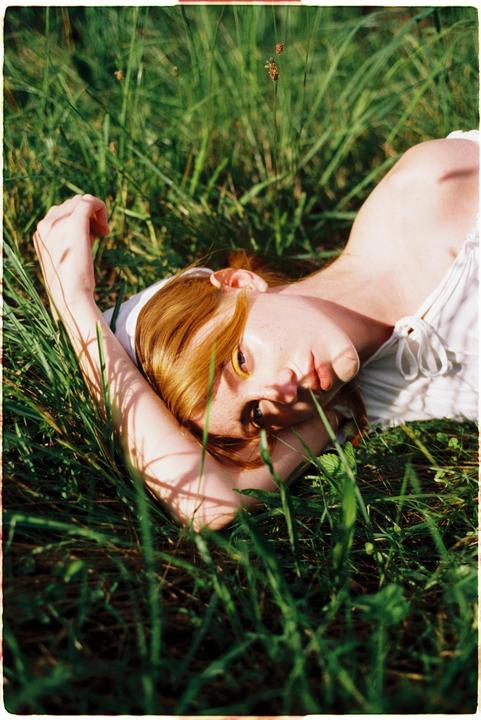 A woman with red hair and freckles is lying in tall green grass, looking up at the viewer.