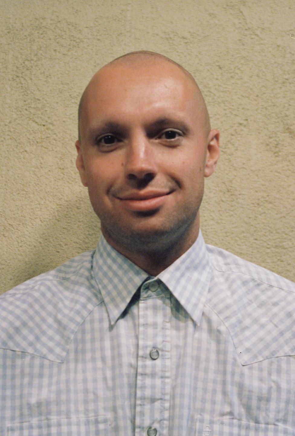 A man with a shaved head and a checkered shirt smiles at the viewer.