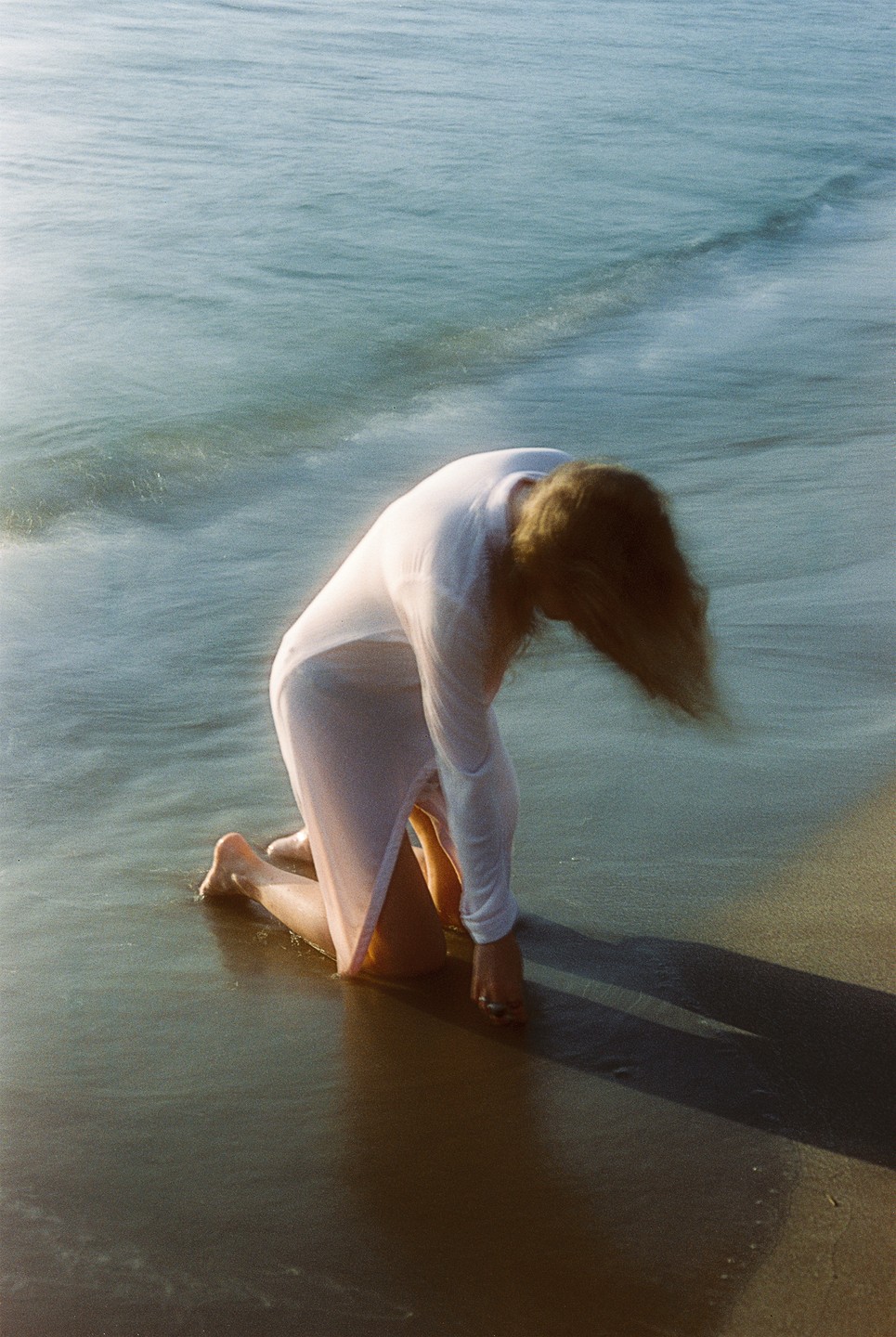 A woman kneels in the shallow water at the edge of a beach, her hair falling forward.