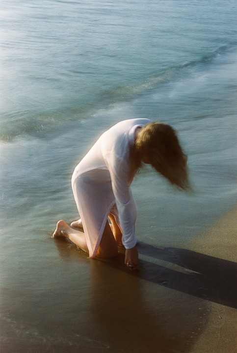 A woman kneels in the shallow water at the edge of a beach, her hair falling forward.