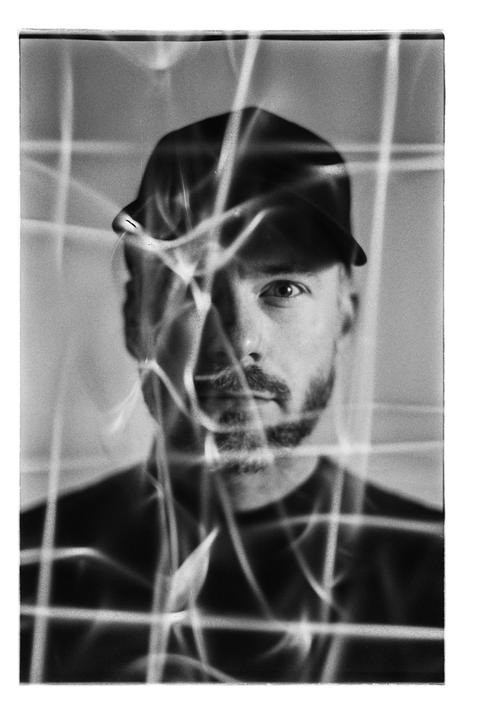 A man wearing a cap looks directly at the viewer through a pattern of bright, abstract lines.