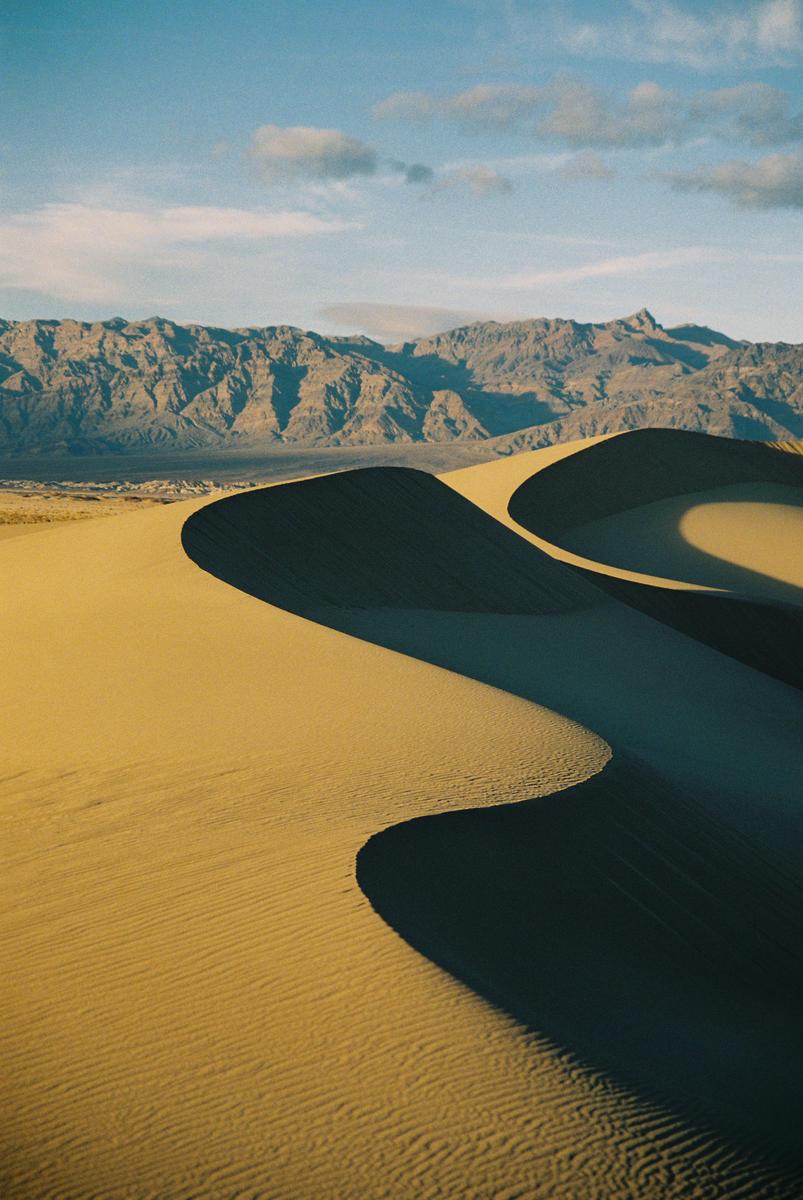 Sand dunes cast long shadows across a desert landscape with mountains in the background under a blue sky.