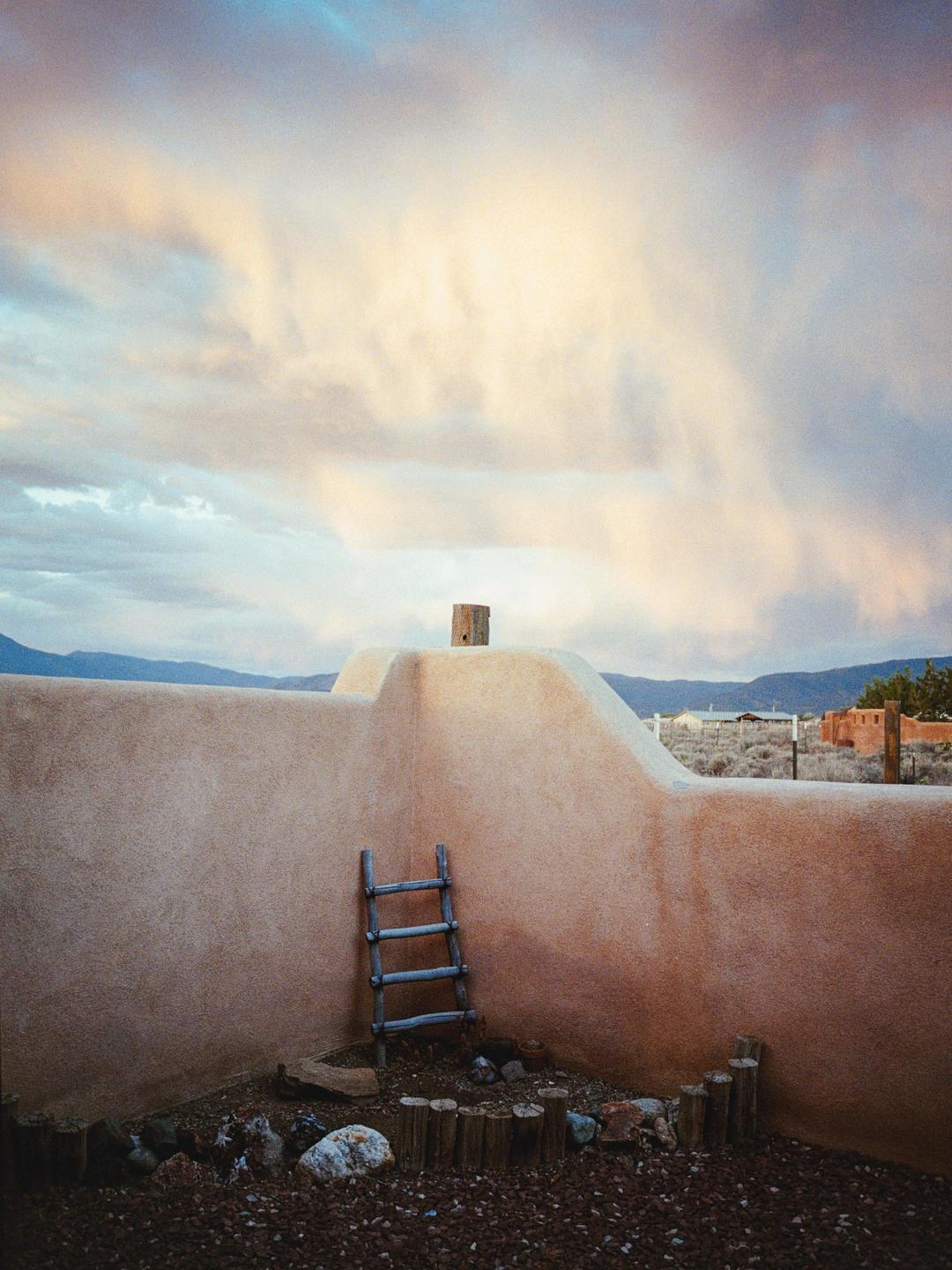 A wooden ladder leans against an adobe wall under a cloudy sky.