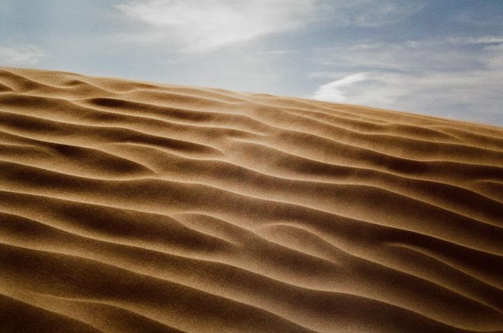 A sand dune with ripples covers the lower two-thirds of the frame, with a blue sky and clouds above.