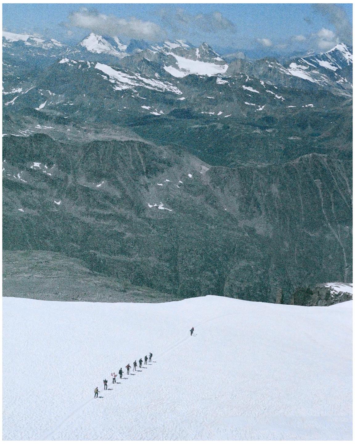 A group of people walk in a line across a snow-covered mountain with a vast mountain range in the background.