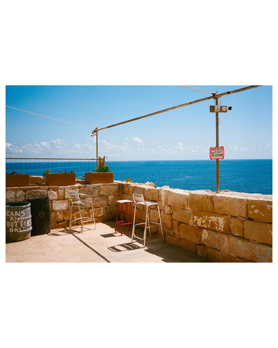 Three bar stools sit on a stone patio overlooking a bright blue sea under a clear sky.