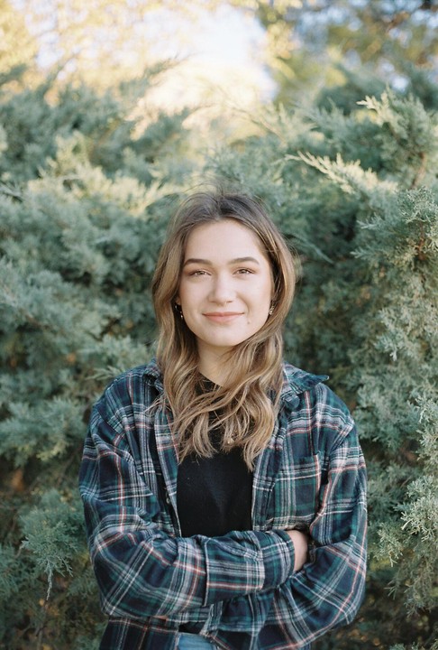 A young woman with long hair and a plaid shirt stands in front of green foliage.