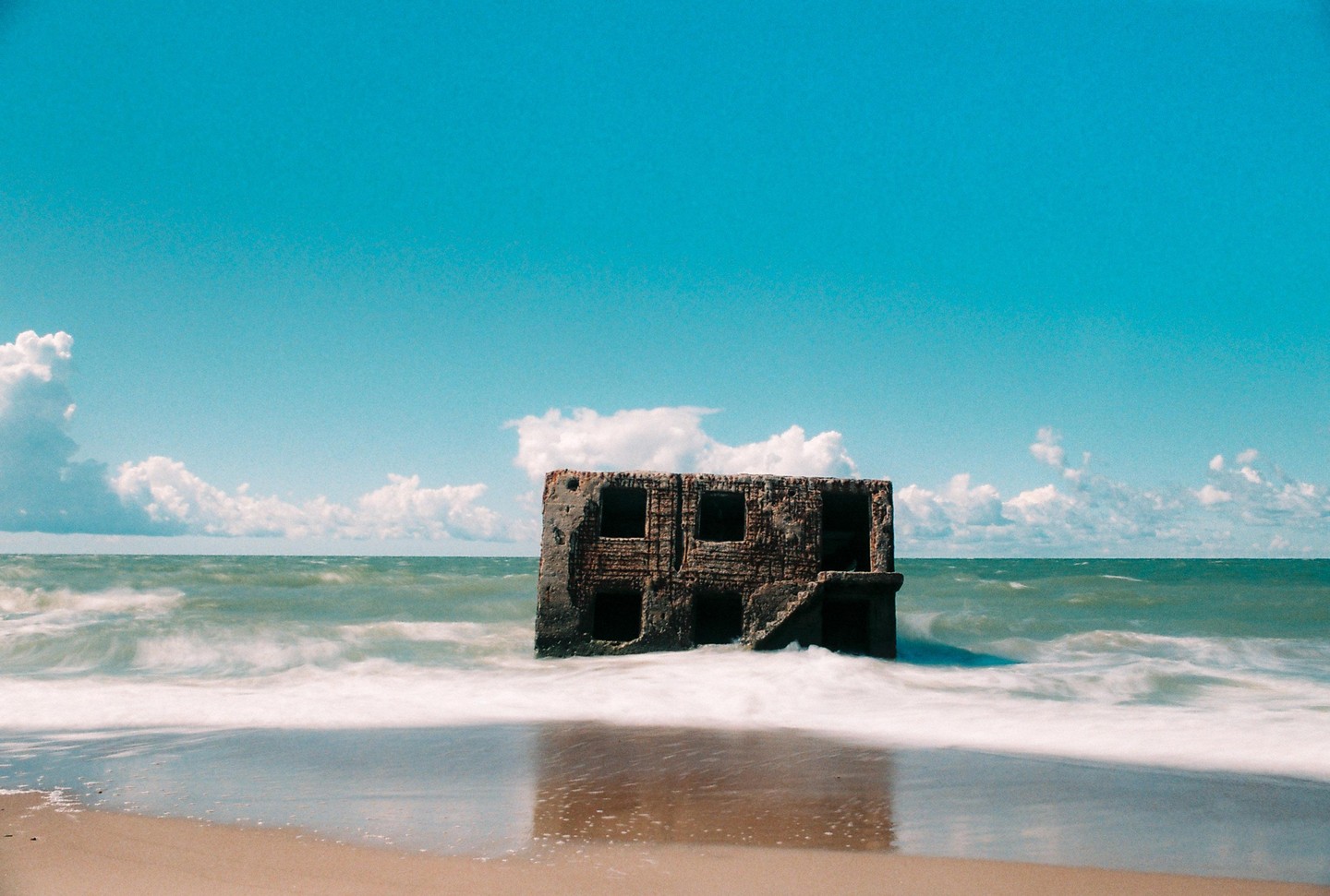 A ruined building stands in the ocean with waves washing onto a sandy beach under a blue sky with clouds.