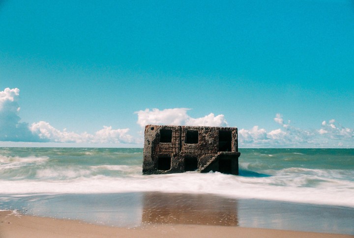 A ruined building stands in the ocean with waves washing onto a sandy beach under a blue sky with clouds.