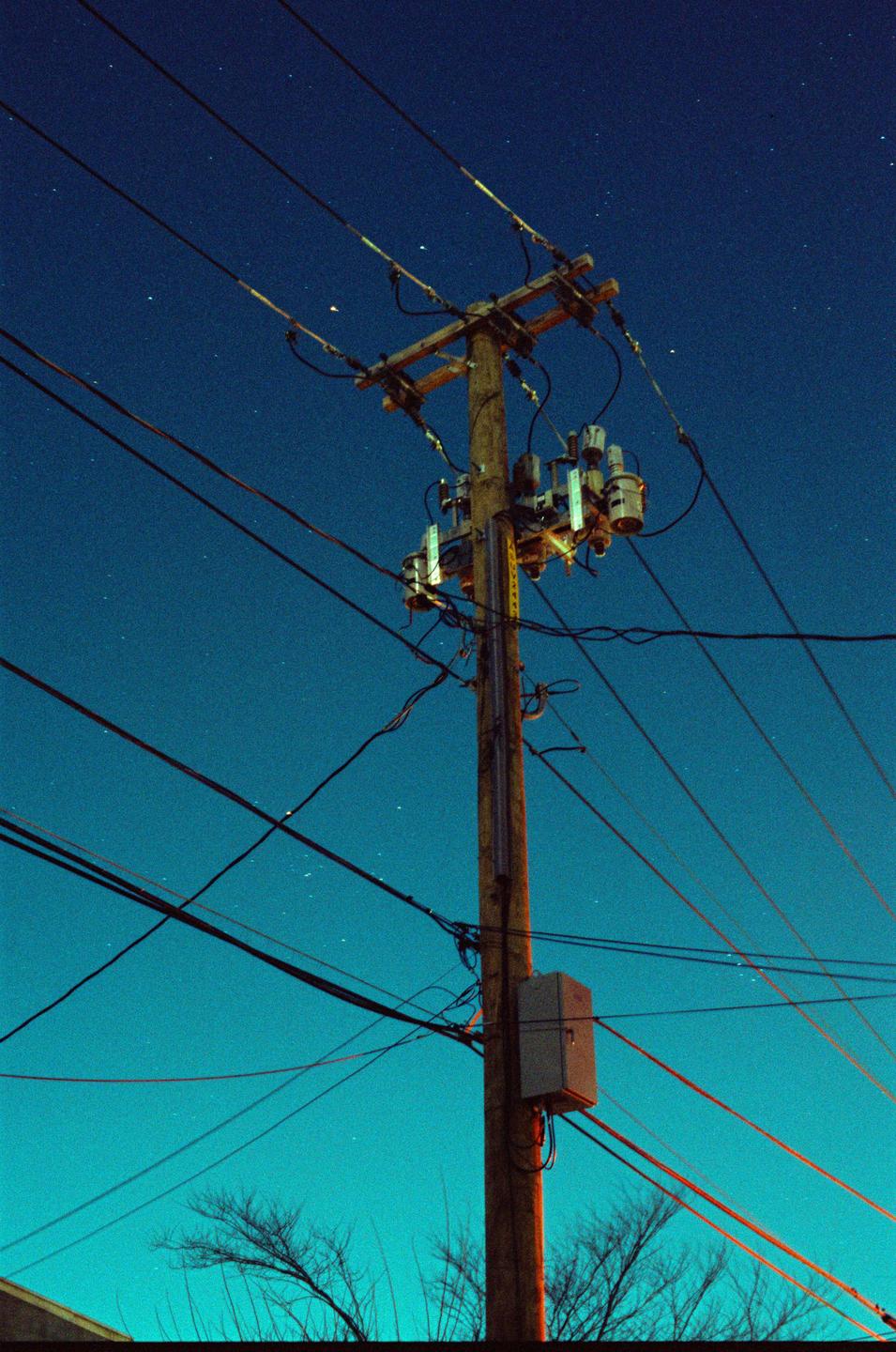 A wooden utility pole with many wires and electrical components stands against a dark blue night sky with visible stars.