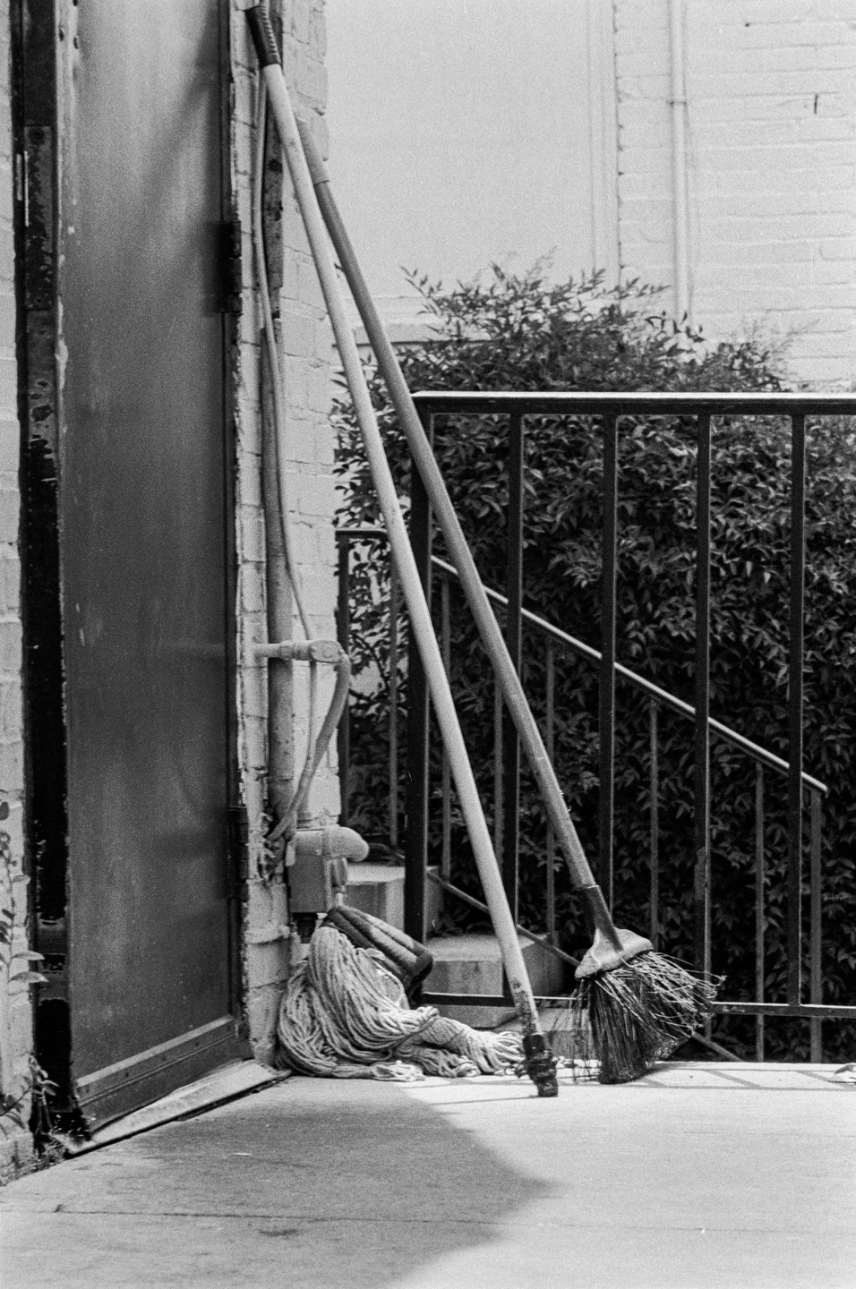 A mop and a broom lean against a building next to a metal railing and bushes.