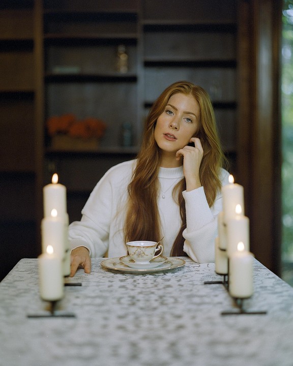 A woman with long red hair sits at a table with lit candles and a teacup.