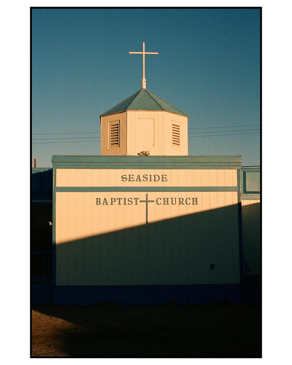 A white church with a teal roof and a cross on top is illuminated by sunlight, casting a diagonal shadow across its facade.