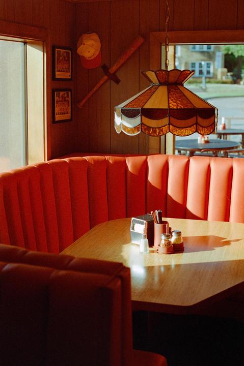 Sunlight illuminates a diner booth with red upholstery and a wooden table set with condiments.