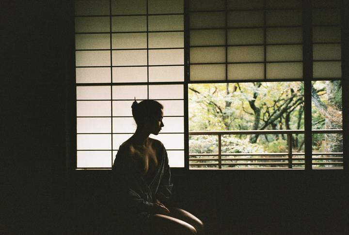 A woman sits in silhouette in front of a window with shoji screens and a view of trees.