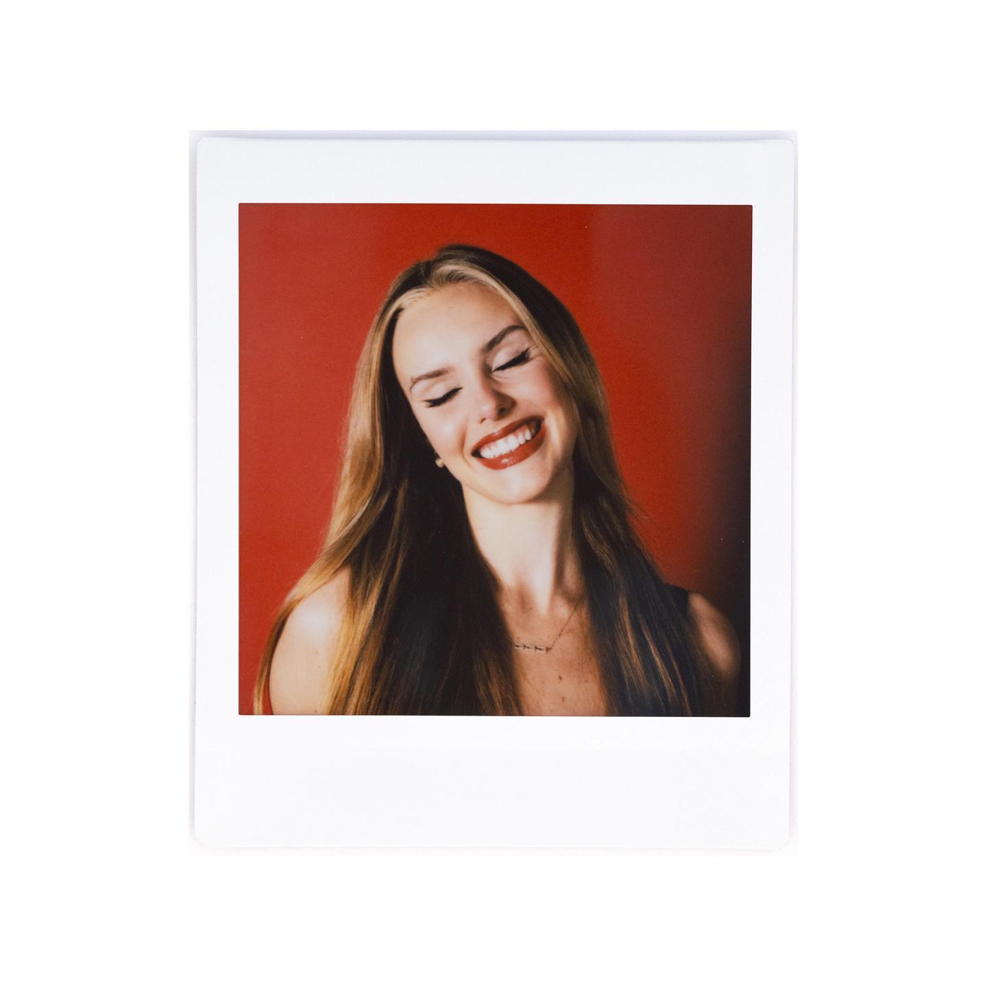 A woman with long hair and red lipstick smiles with her eyes closed against a red background.