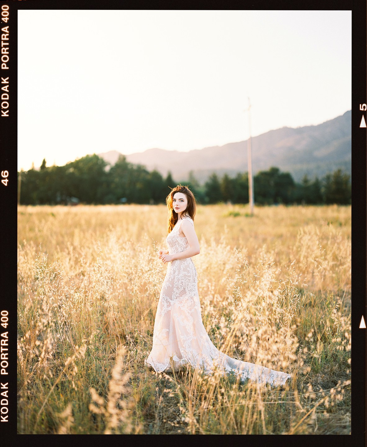 A woman in a long, light-colored dress stands in a field of tall grass with mountains in the background.