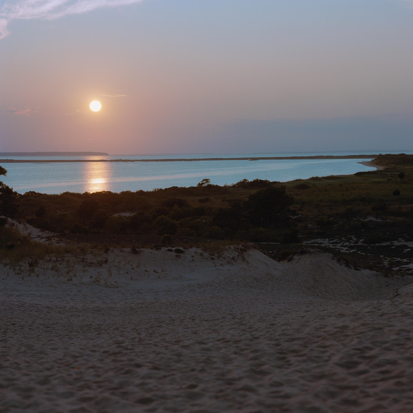 The sun sets over a body of water with sand dunes and vegetation in the foreground.