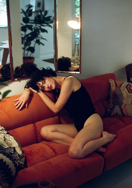 A woman with curly hair in a black bodysuit is reclining on an orange sofa, looking towards the viewer.