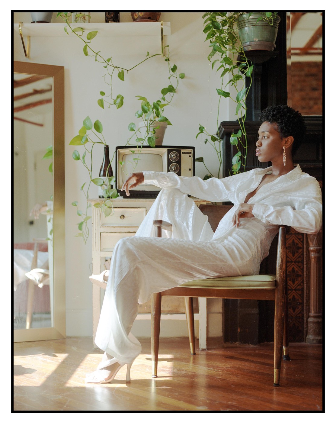 A woman in a white outfit sits on a wooden chair in a room with plants and a vintage television.
