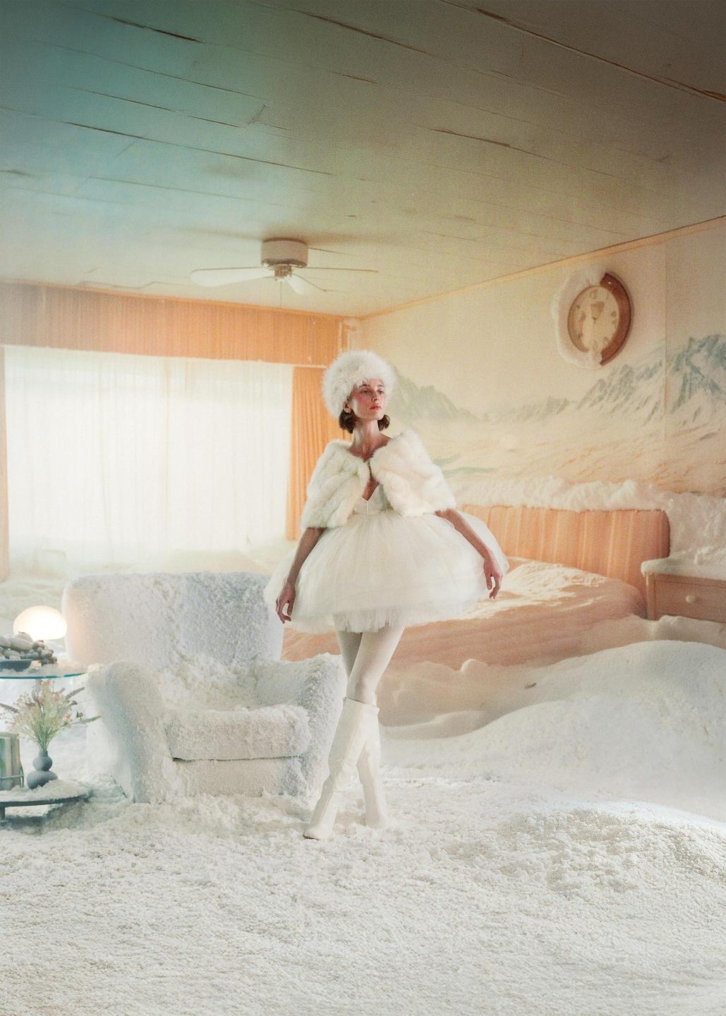A woman in a white fur hat and cape stands in a room covered in white powder, with a mountain mural on the wall.
