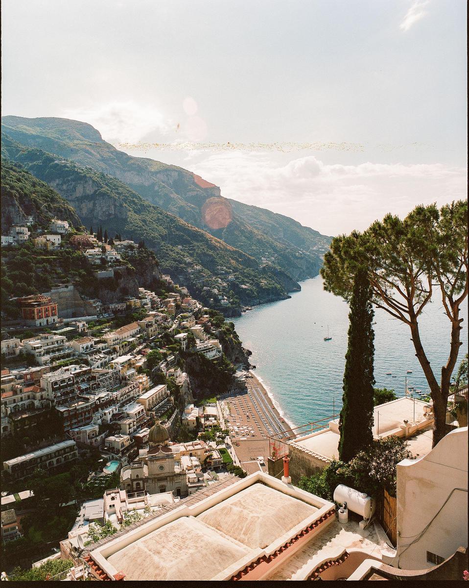 A hillside town with many buildings and a beach is visible next to a body of water with mountains in the background.