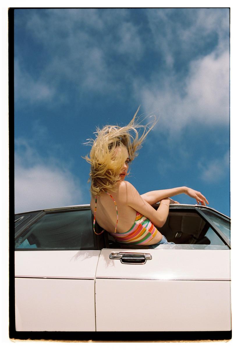A woman with blonde hair blowing in the wind leans out of a white car against a blue sky with clouds.