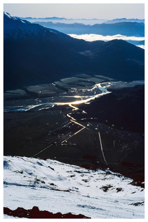 A winding river reflects sunlight through a valley surrounded by mountains, with a snowy slope in the foreground.