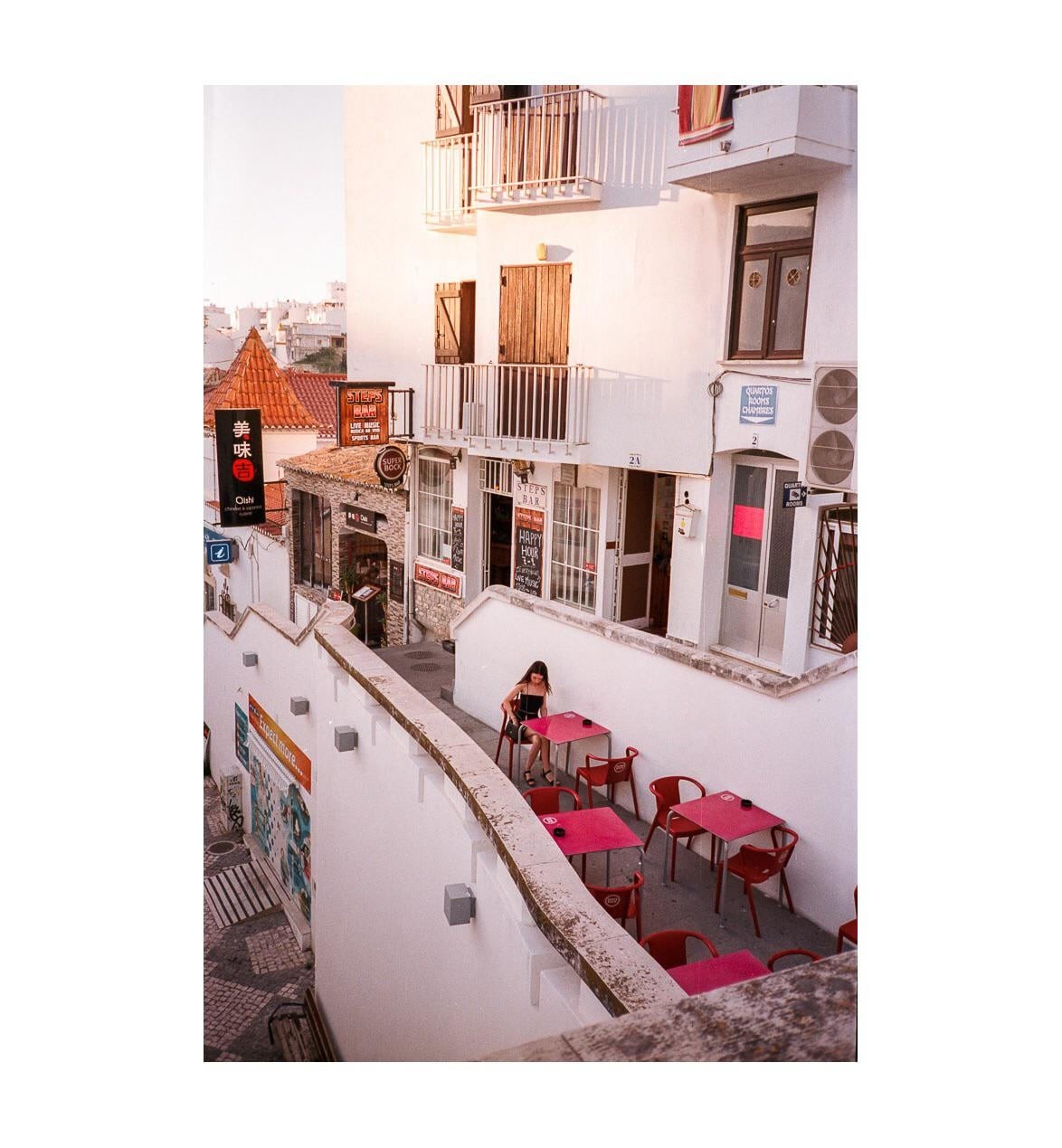 A woman sits alone at a red table on a terraced street with white buildings and shops.