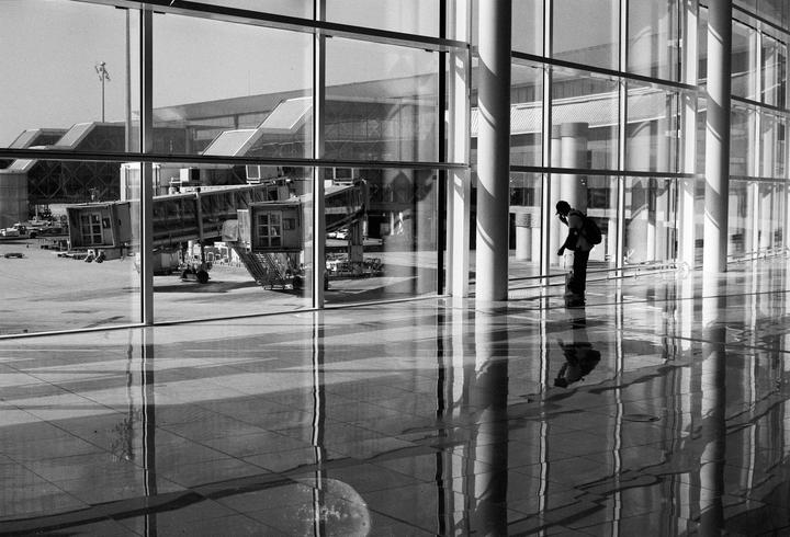 A person wearing a backpack stands in a modern building with large windows overlooking an airport tarmac with jet bridges.