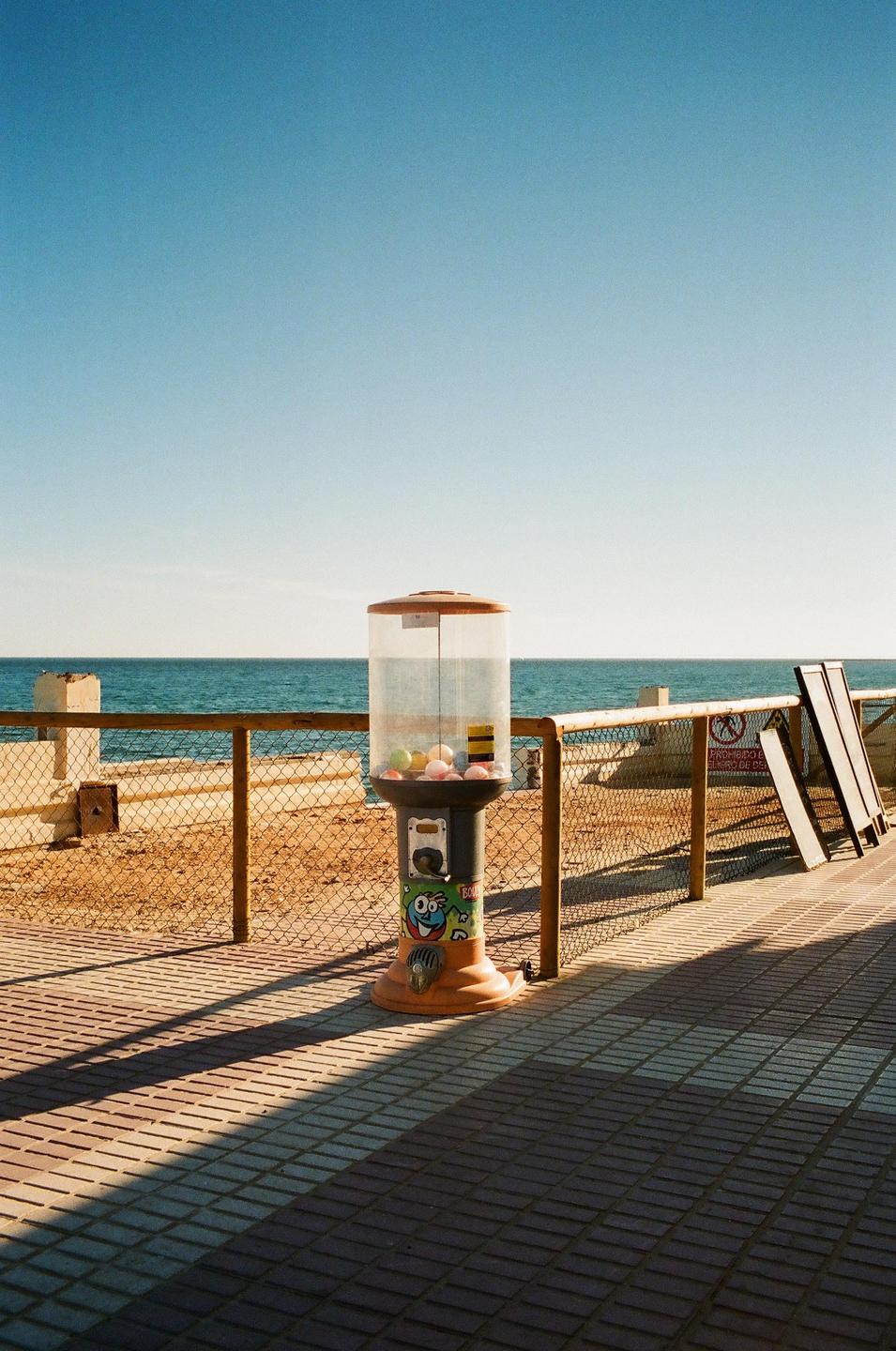 A gumball machine stands on a tiled promenade next to a fence, with the ocean and a clear sky in the background.