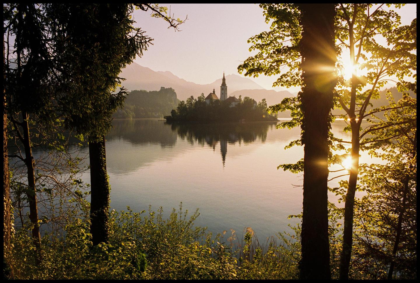 A church on an island in a lake is framed by trees with mountains in the background at sunset.