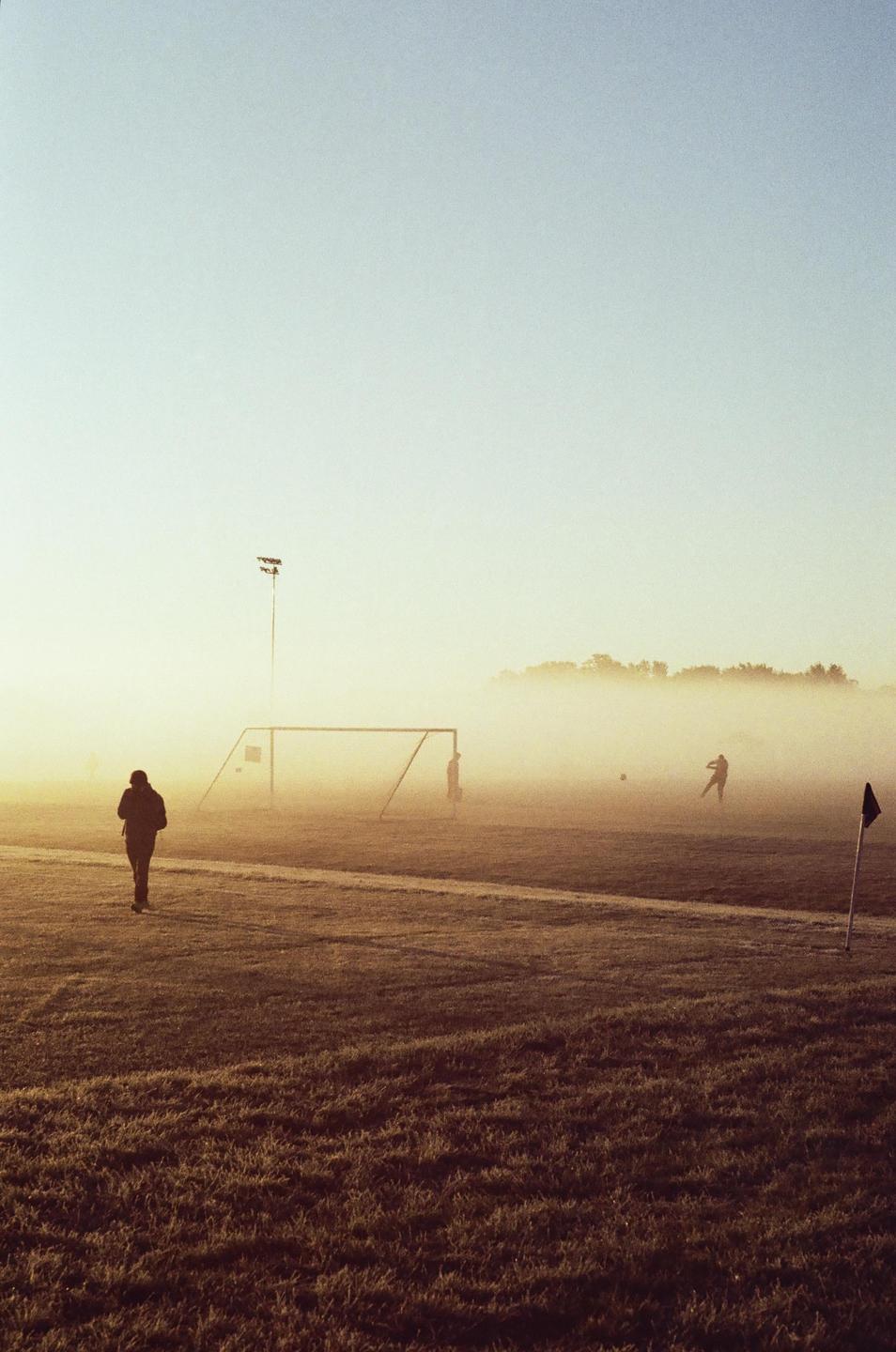A person walks across a foggy soccer field with a goal and other people in the distance.