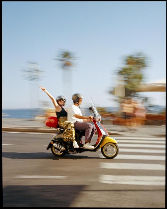 A couple rides a scooter on a road next to the ocean.